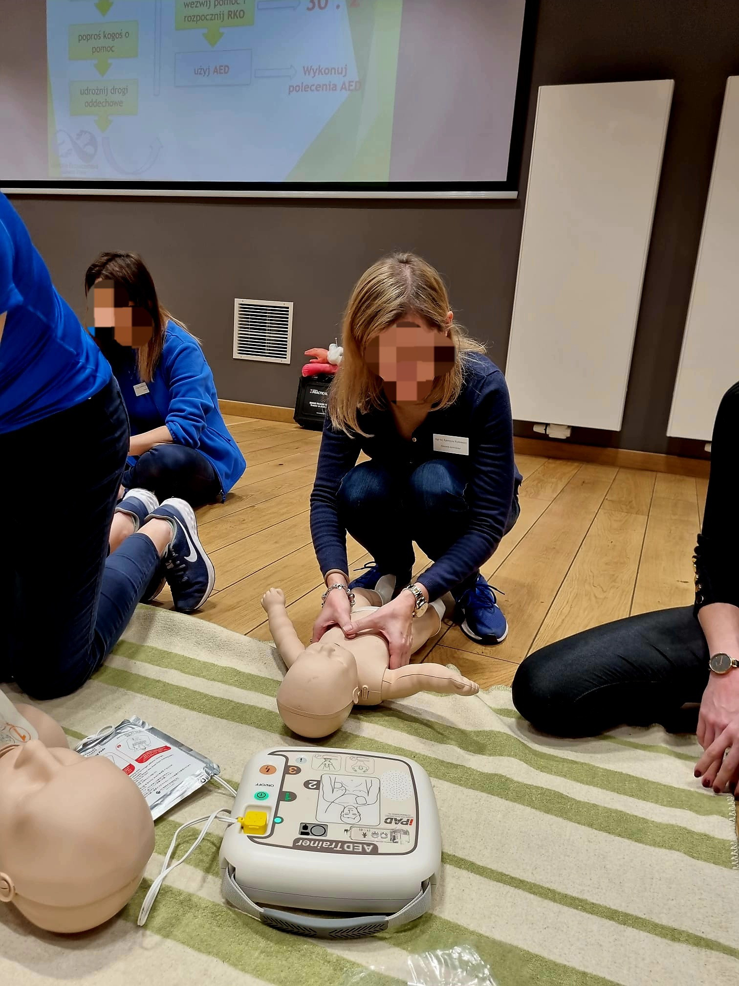 Instruktor demonstruje resuscytację krążeniowo-oddechową niemowlęcia na manekinie podczas szkolenia. Na pierwszym planie AED Trainer, w tle slajd z algorytmem postępowania.