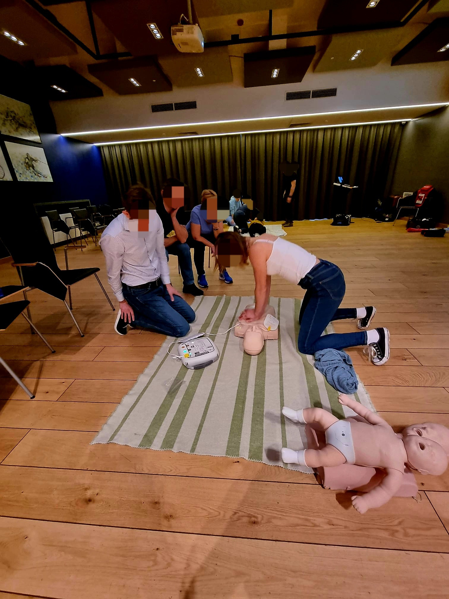 Sala szkoleniowa z uczestnikami ćwiczącymi resuscytację krążeniowo-oddechową na manekinach niemowląt i osoby dorosłej, z użyciem defibrylatora.