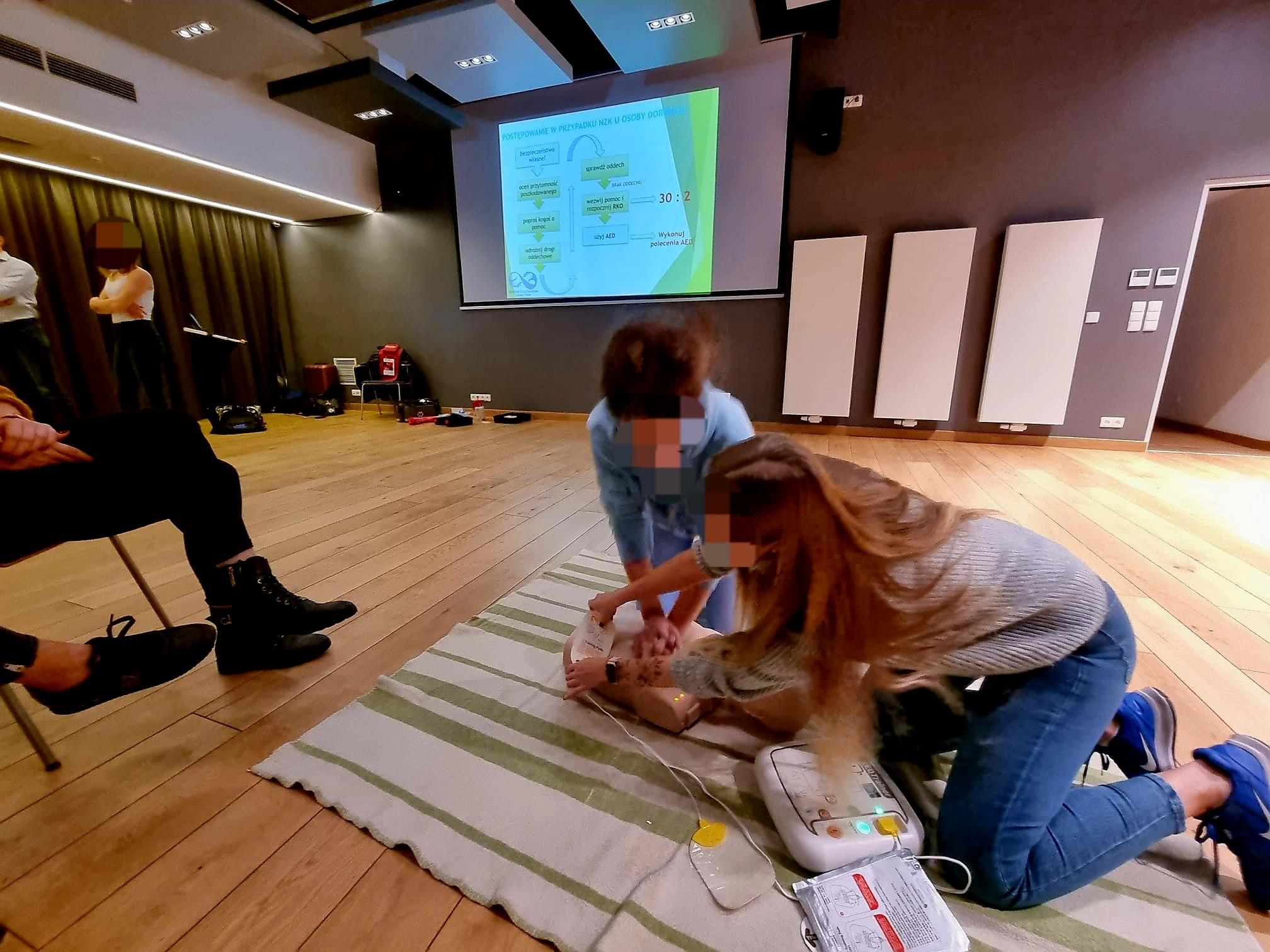 Sala szkoleniowa z drewnianą podłogą; dwie osoby ćwiczą resuscytację krążeniowo-oddechową na manekinie, używając defibrylatora AED; na ścianie wyświetlany schemat postępowania w przypadku NZK.
