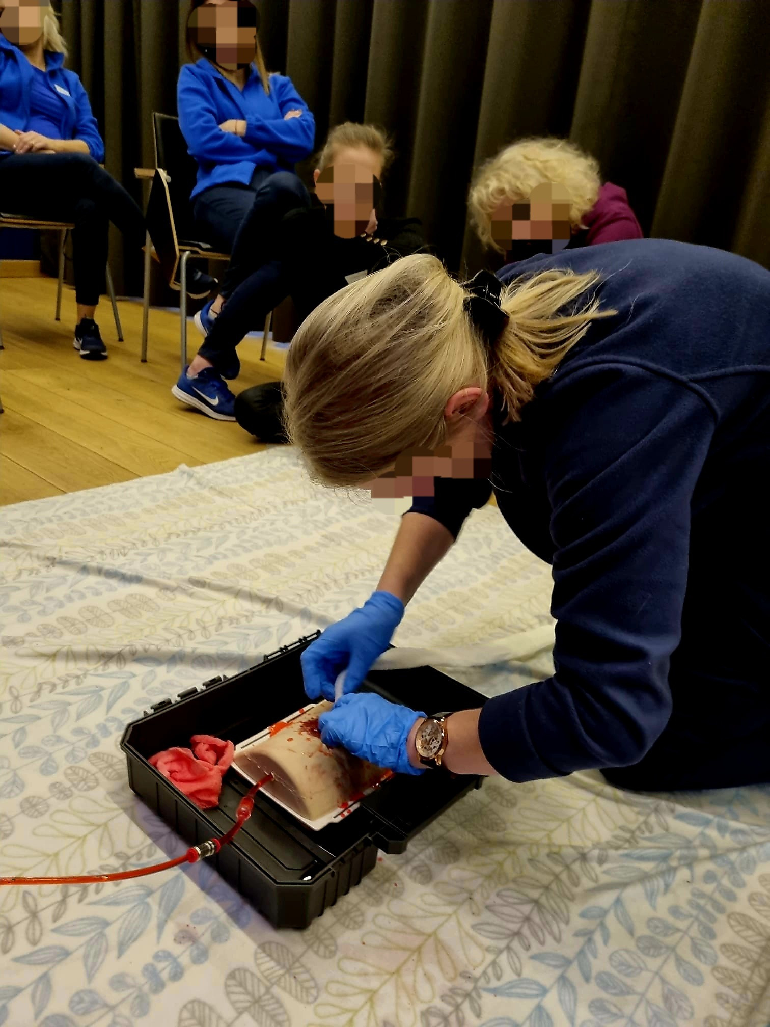 Instruktor w niebieskich rękawiczkach demonstruje na fantomie w czarnej skrzynce, jak zatamować krwotok, w tle obserwujący kursanci.