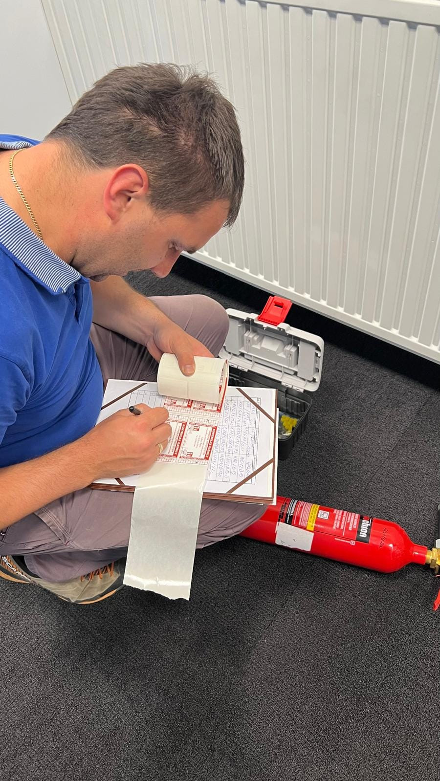 Mężczyzna siedzi na podłodze obok czerwonej gaśnicy, wypełniając etykiety kontrolne z rolki naklejek, obok skrzynka z narzędziami i grzejnik.