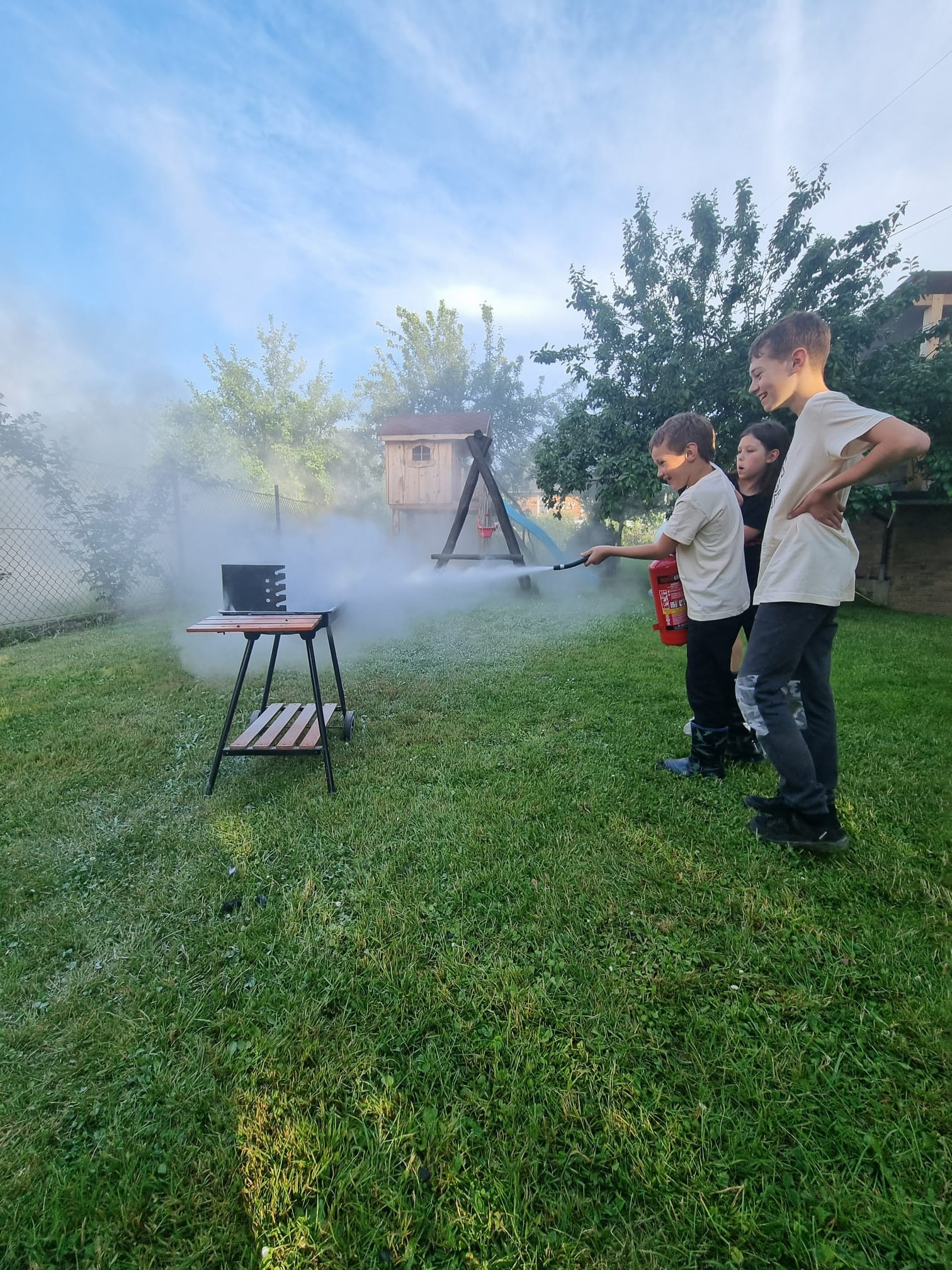Troje dzieci na trawniku ćwiczy gaszenie pożaru na grillu za pomocą czerwonej gaśnicy, w tle widoczny domek na drzewie i błękitne niebo.