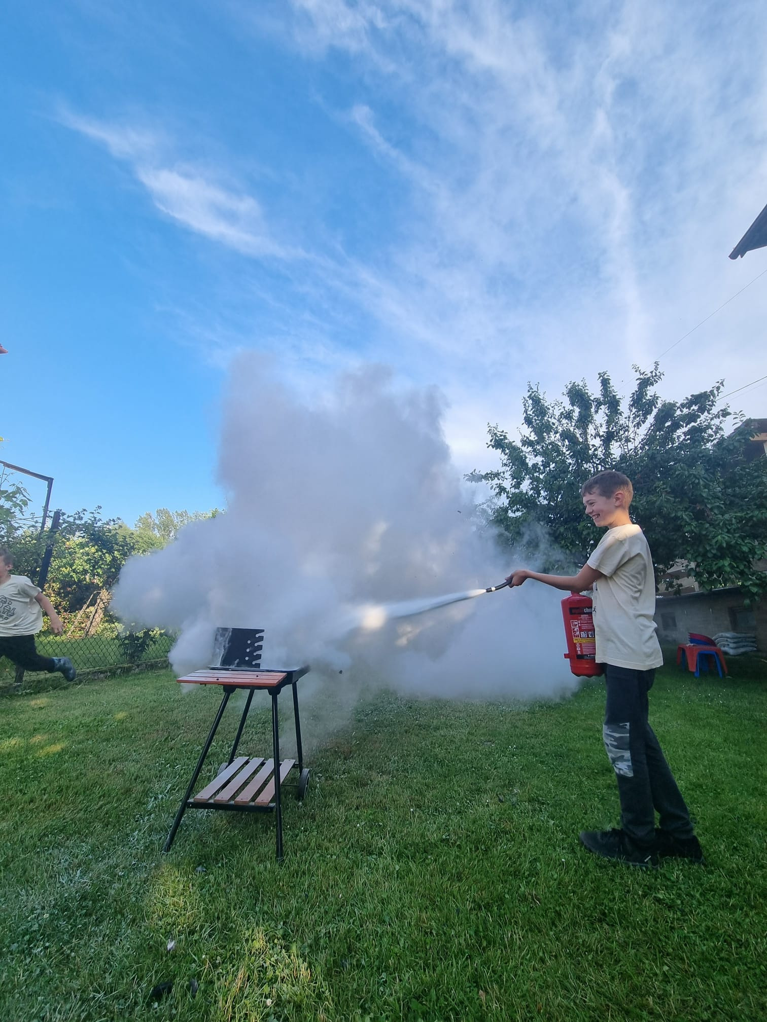 Chłopiec w jasnej koszulce i ciemnych spodniach z uśmiechem gasi pożar grilla za pomocą czerwonej gaśnicy, podczas gdy drugi chłopiec biegnie w oddali, na tle zielonego trawnika i błękitnego nieba.