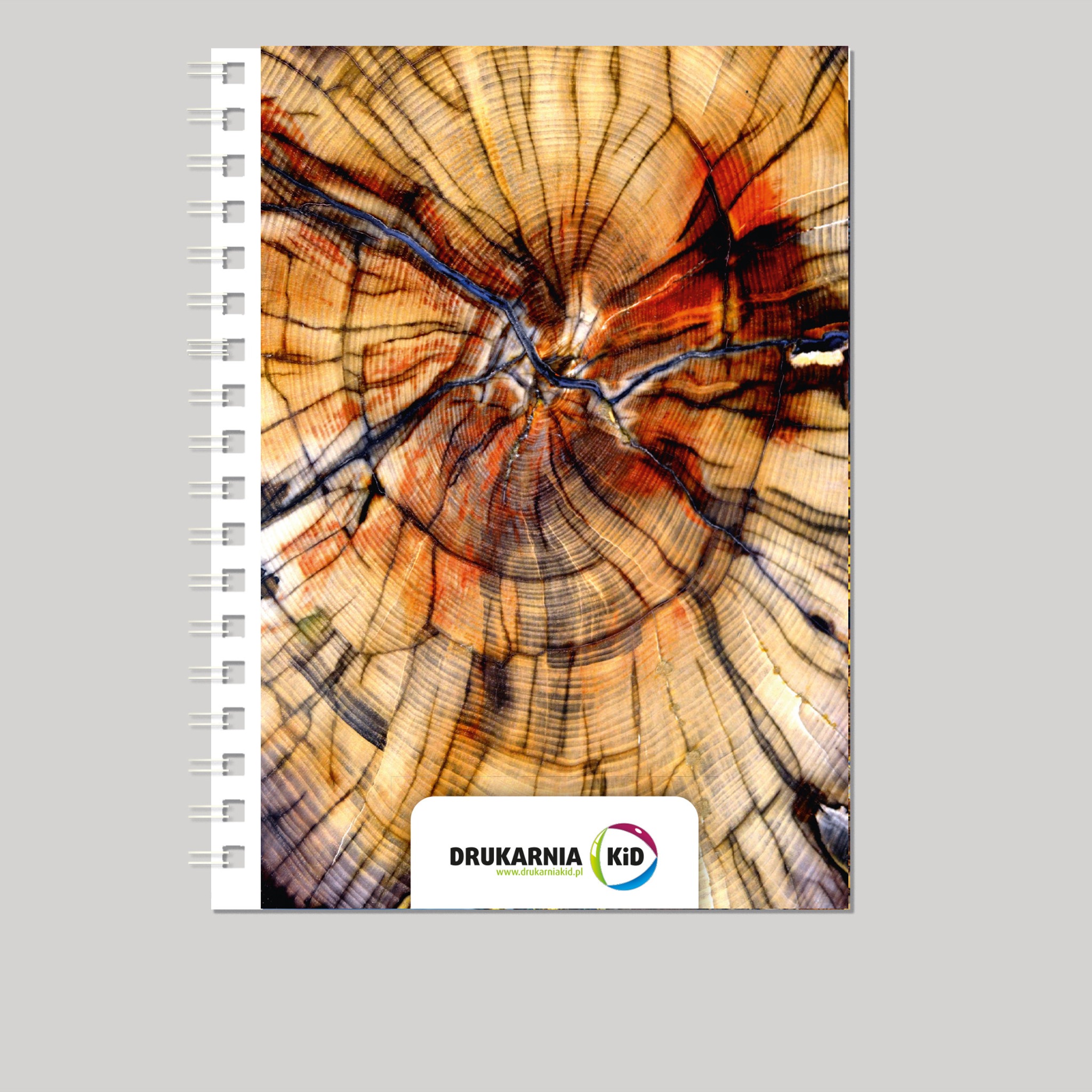 Notatnik spiralny z okładką przedstawiającą przekrój pnia drzewa z wyraźnymi słojami i logo drukarni w dolnej części.