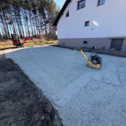 Przygotowany teren pod budowę: ubity żwir, widoczny zagęszczarką, w tle dom jednorodzinny i minikoparka. Grunt wyrównany, gotowy do dalszych prac.