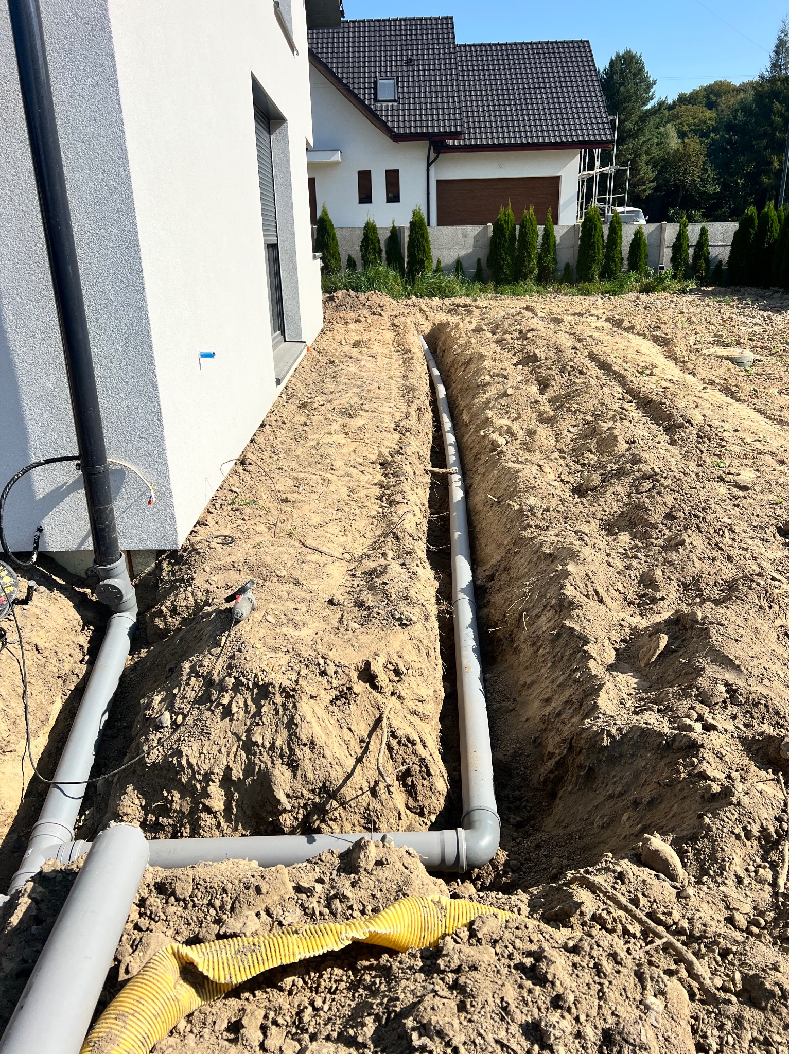 Wykop wzdłuż ściany budynku z ułożonymi rurami drenażowymi, widoczne połączenia i żółta rura karbowana, w tle drugi budynek i tuje.