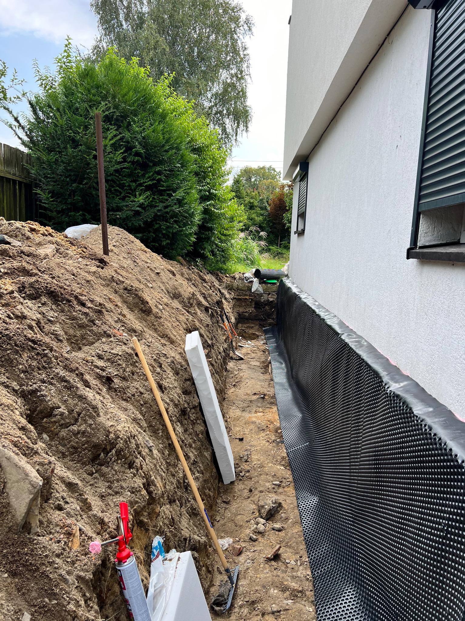 Odkryte fundamenty domu z nałożoną czarną folią kubełkową, zabezpieczającą przed wilgocią, obok wykopu leżą płyty styropianowe i narzędzia, ziemia usypana w pryzmę.