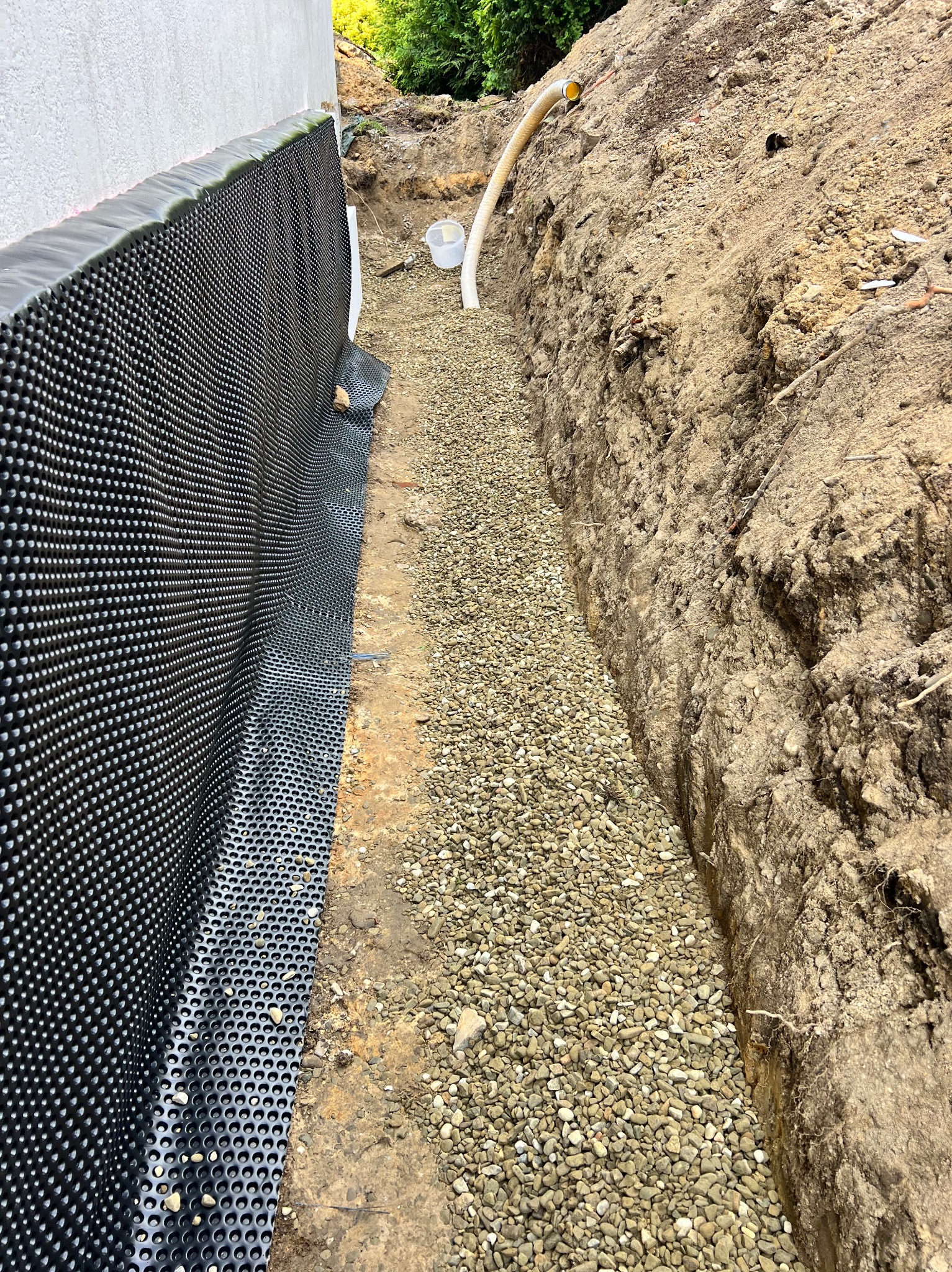 Czarna folia kubełkowa przy ścianie fundamentowej, drenaż z drobnych kamieni w wykopie, widoczna rura drenażowa i wiaderko.
