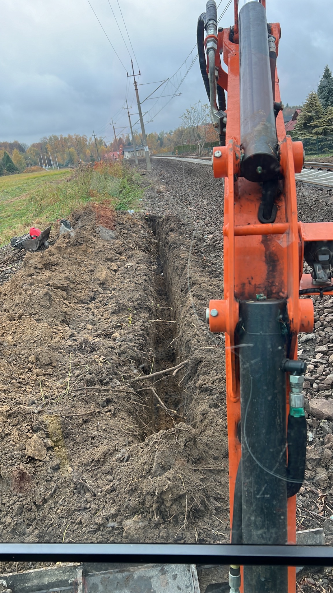 Wąski wykop liniowy w ziemi, wykonany wzdłuż torów kolejowych, z widocznym pomarańczowym ramieniem koparki i elementami krajobrazu w tle pod pochmurnym niebem.