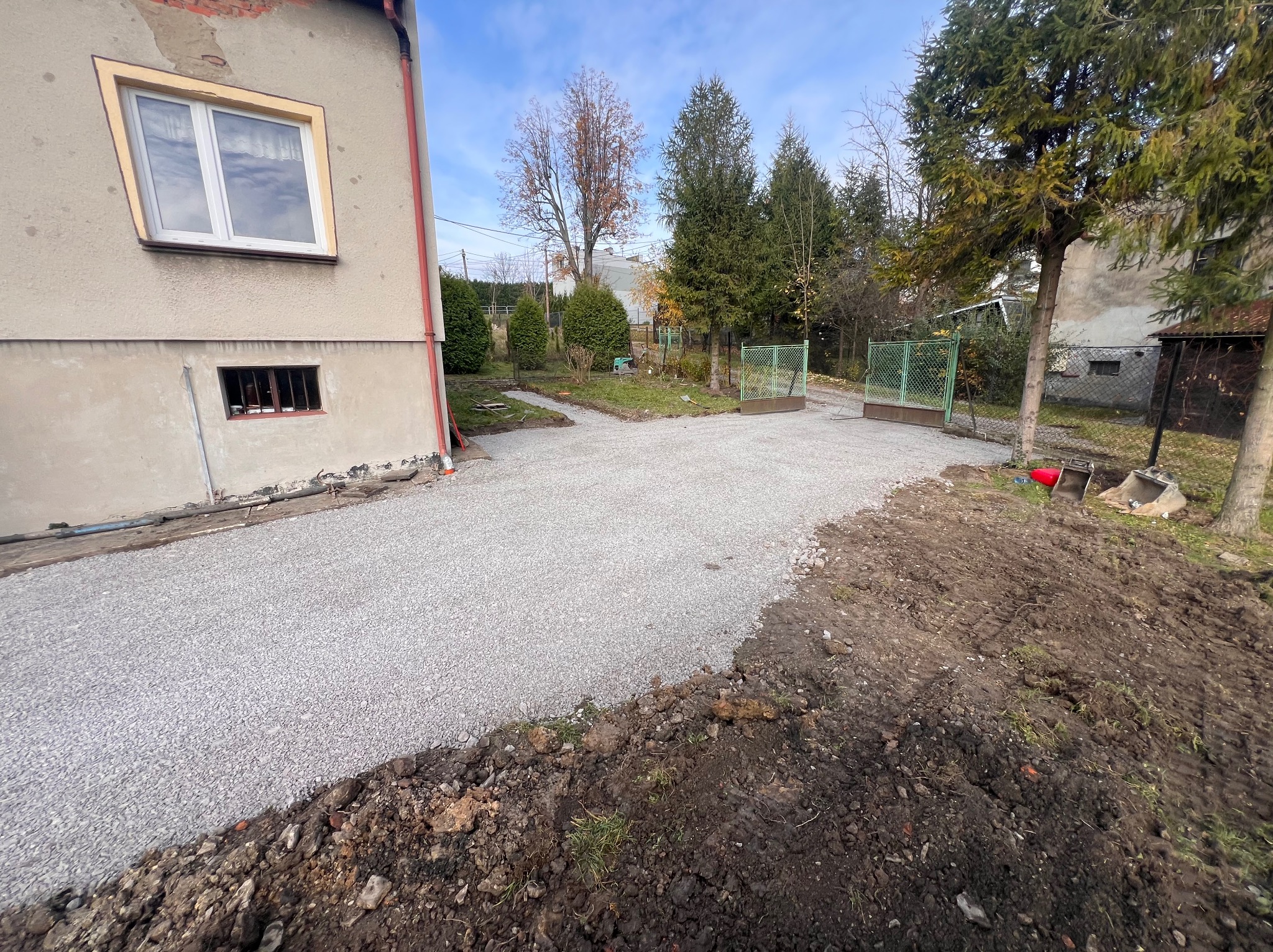 Świeżo utwardzona żwirowa droga dojazdowa przed domem z widoczną ziemią po bokach i zielonym ogrodzeniem w tle.