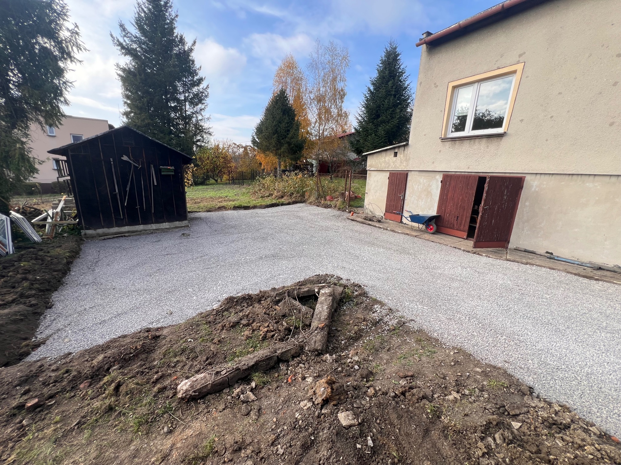 Przygotowany teren pod budowę z wysypanym drobnym żwirem, widoczny budynek gospodarczy i dom w tle, częściowo odsłonięta ziemia z fragmentami betonu na pierwszym planie.