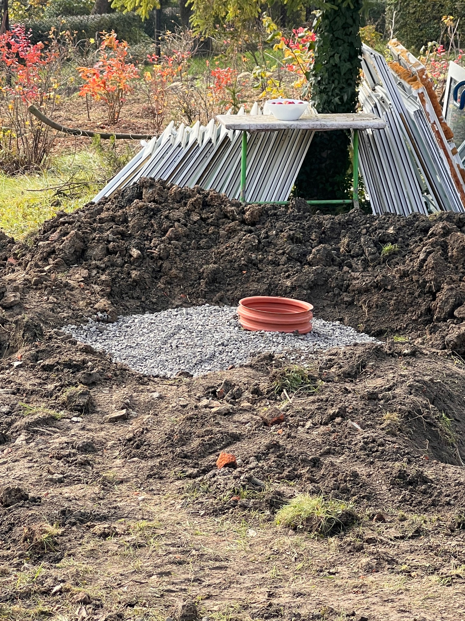 Wykop z warstwą drenażową z kruszywa i rurą drenarską, przygotowany pod fundament w ogrodzie z jesiennymi krzewami w tle.