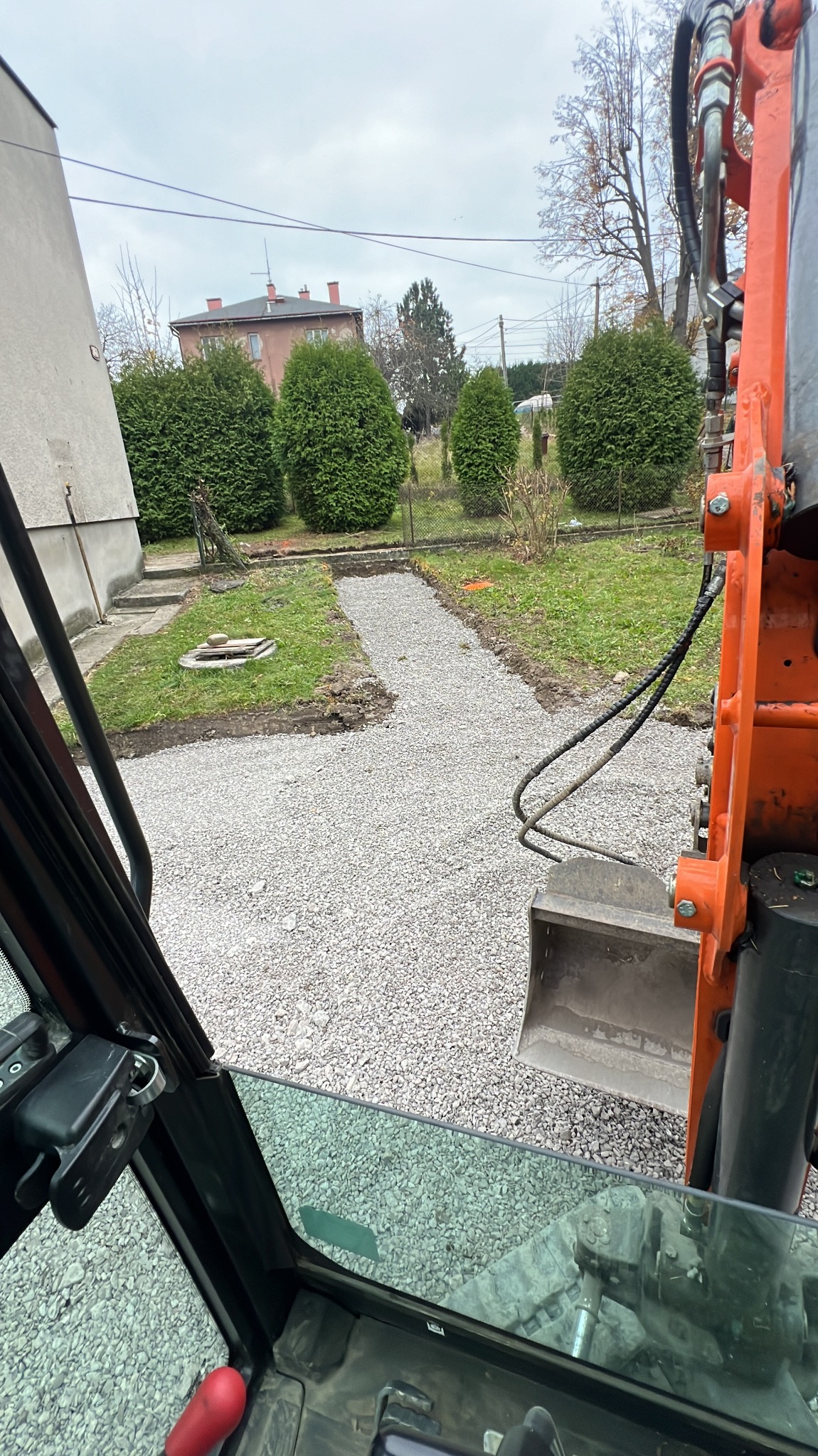 Widok z kabiny pomarańczowej koparki na ścieżkę z drobnego żwiru prowadzącą przez trawnik, z tujami i budynkiem w tle, w pochmurny dzień.