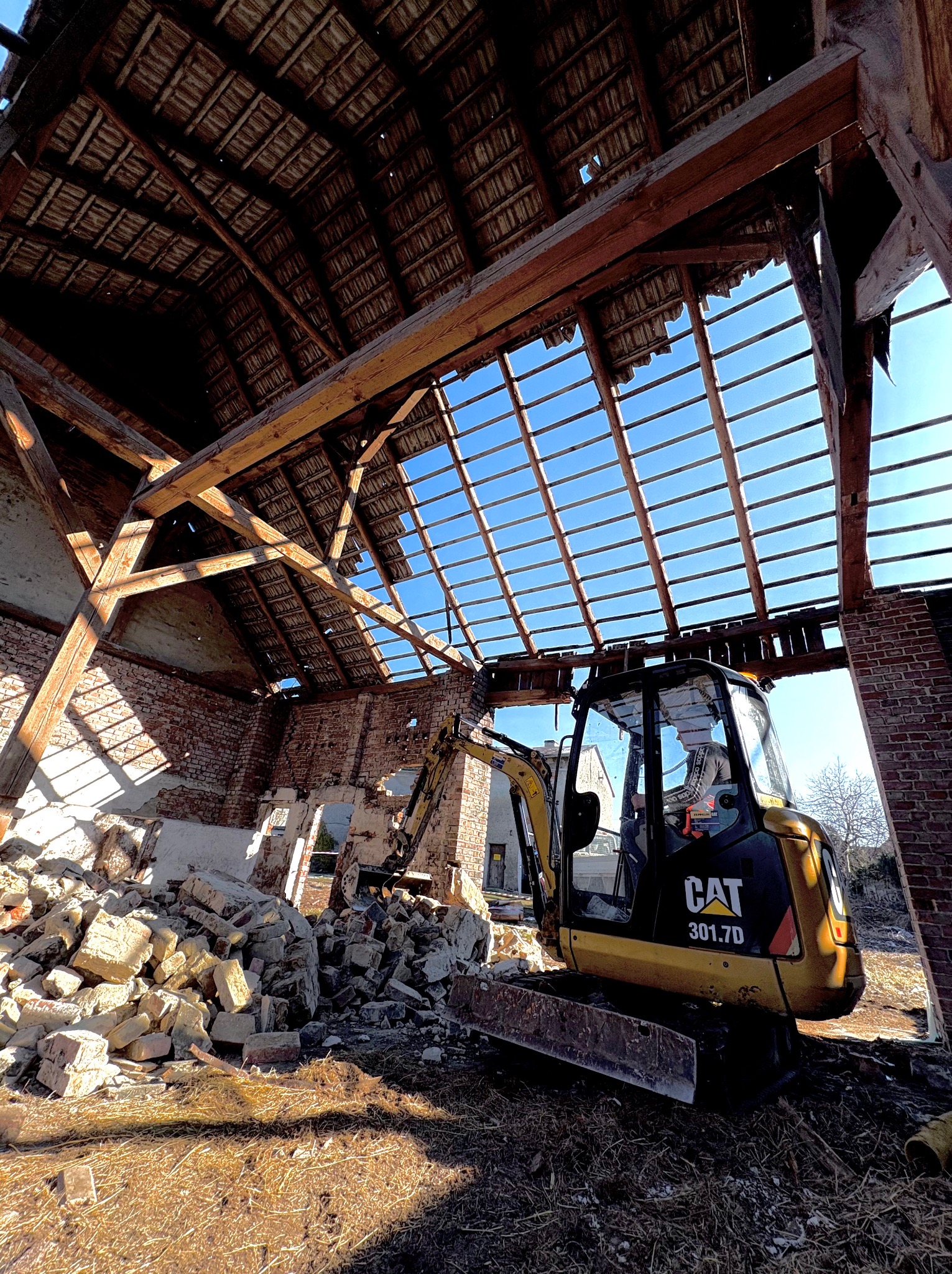 Żółta minikoparka CAT 301.7D wewnątrz zrujnowanej, ceglanej stodoły z częściowo zawalonym, drewnianym dachem i stertą gruzu.