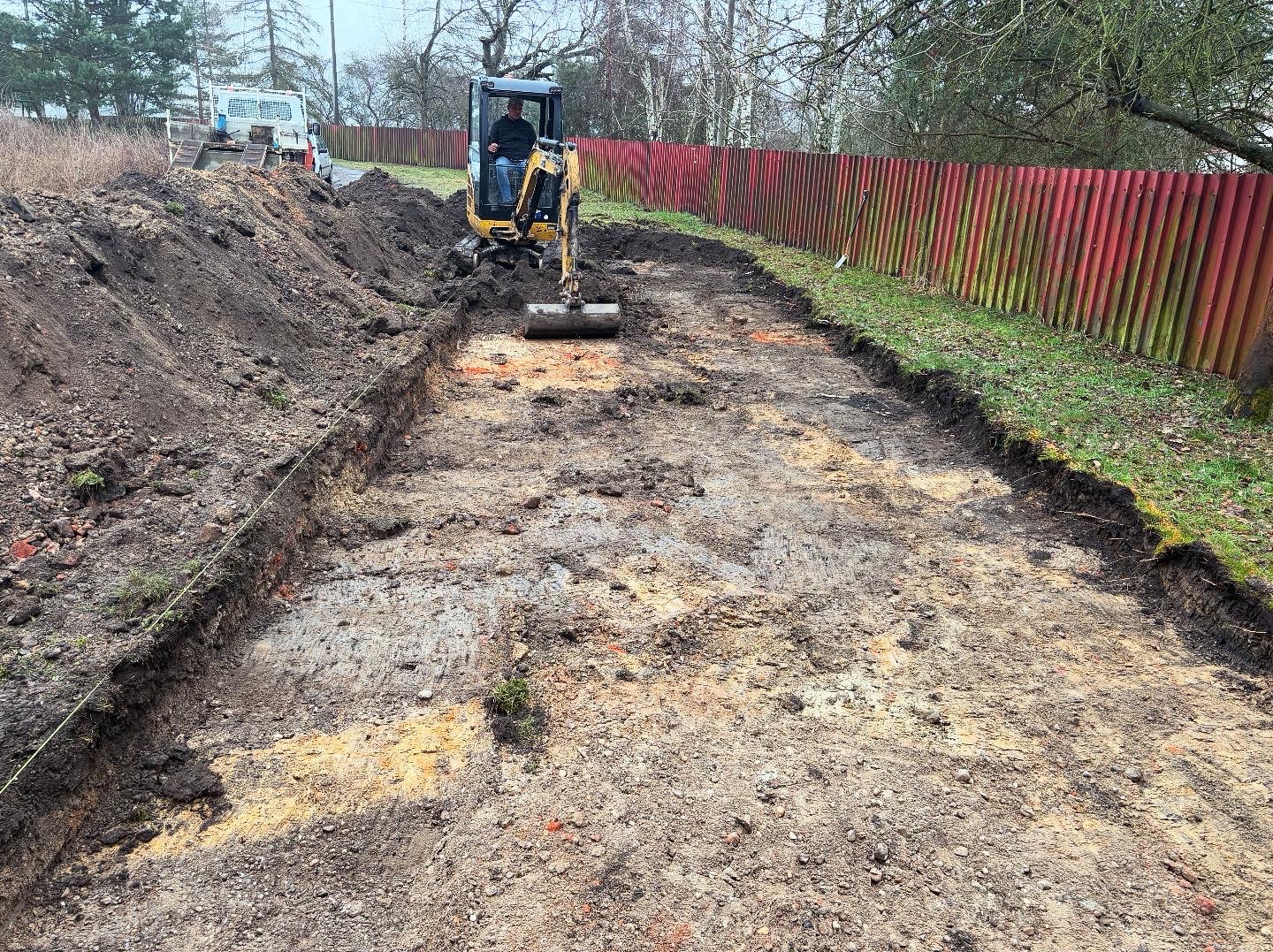Żółta mini koparka pracująca w wykopie pod przyszłą drogę, widoczna czerwona metalowa bariera w tle, obok wykopu leży pryzma ziemi.