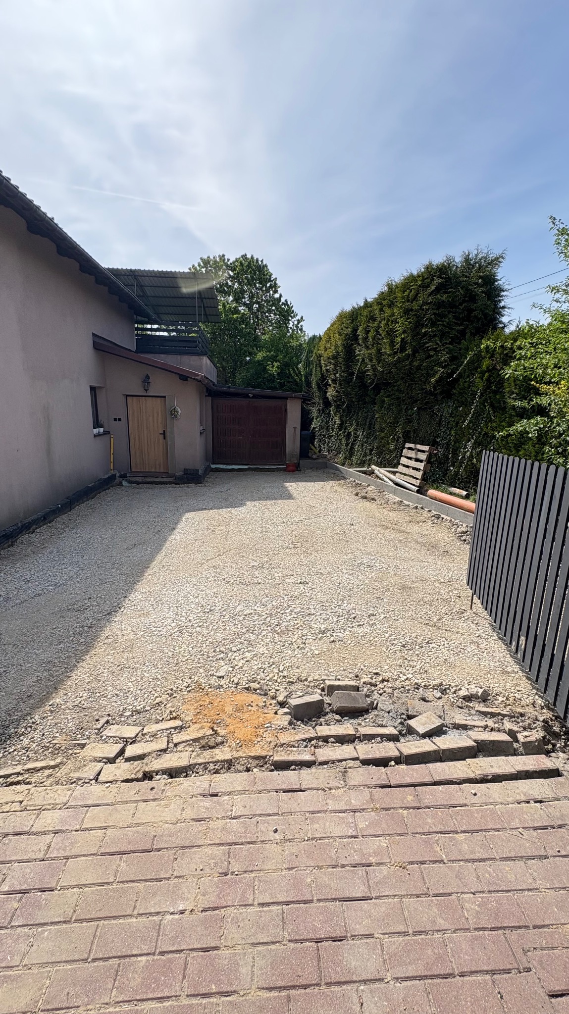 Przygotowanie podłoża z kruszywa na podjeździe przed domem, widoczne obrzeża i częściowo rozebrana kostka brukowa. Prace ziemne w słoneczny dzień.