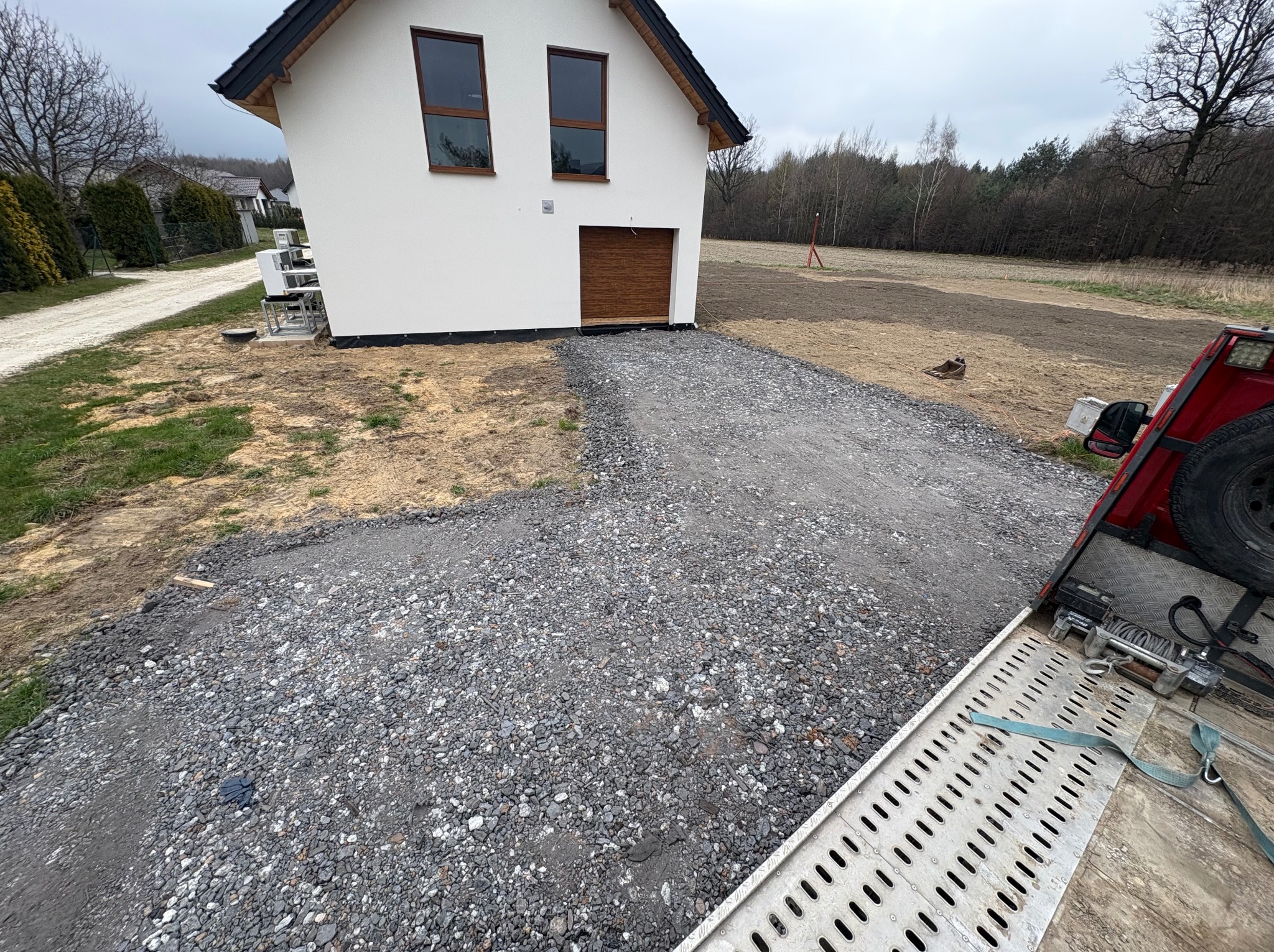 Nowy dom z utwardzoną żwirową drogą dojazdową, widok z rampy samochodu. Świeżo przygotowany teren pod trawnik. Szara, pochmurna pogoda.