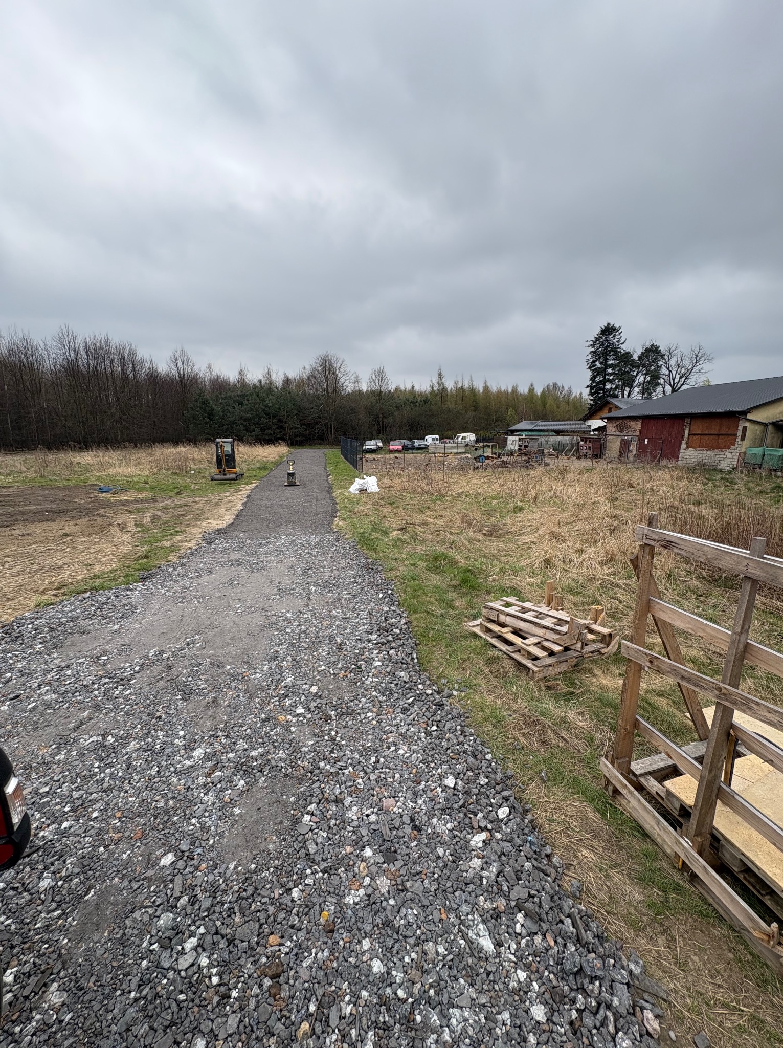 Utwardzona, szara, kamienista droga prowadzi przez pole w kierunku lasu. Na poboczu mini koparka i palety. Pochmurne niebo.