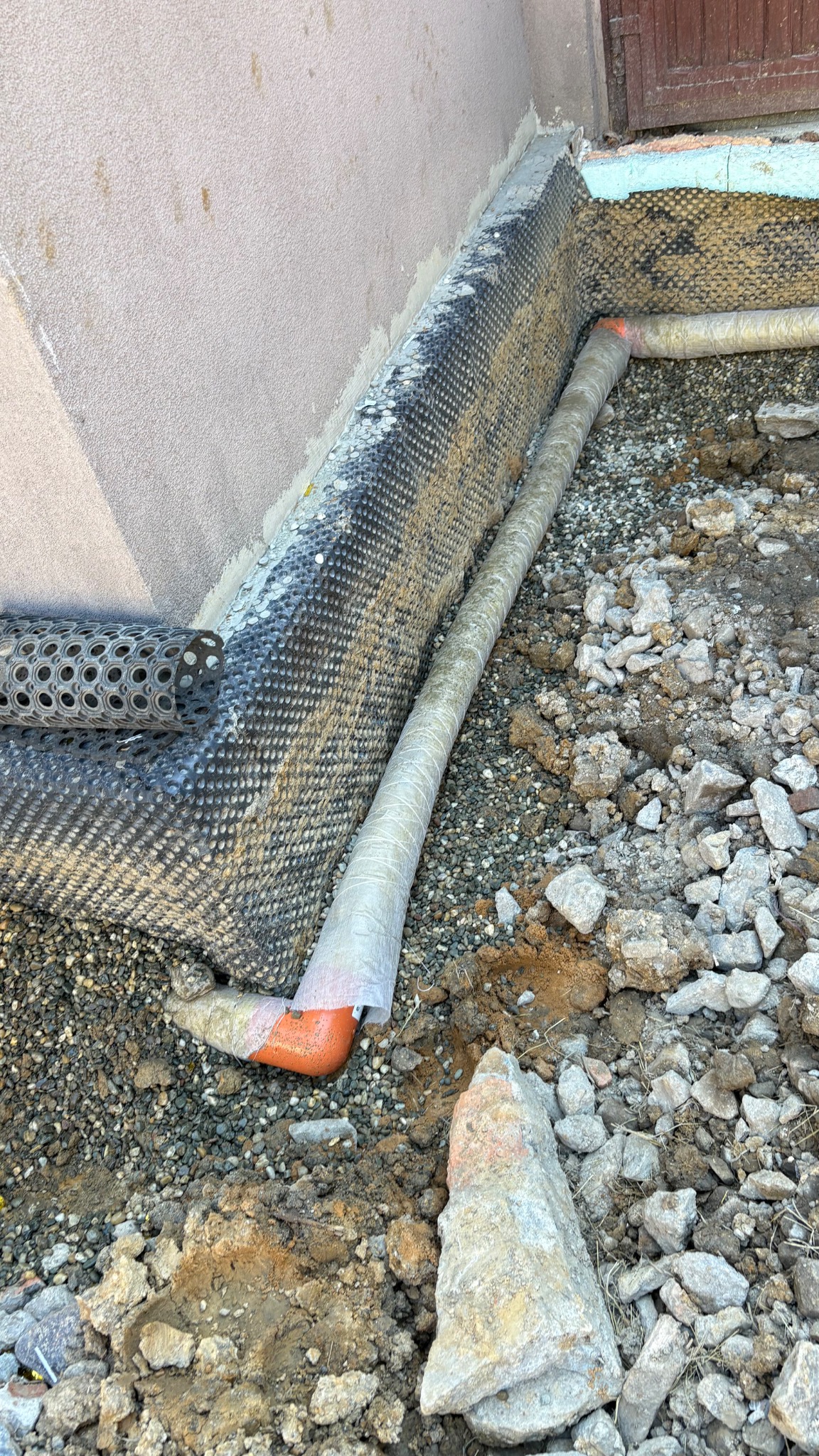 Fundament zabezpieczony folią kubełkową i rurą drenarską, widoczna warstwa żwiru. Etap prac izolacyjnych przy budynku, z bliska.