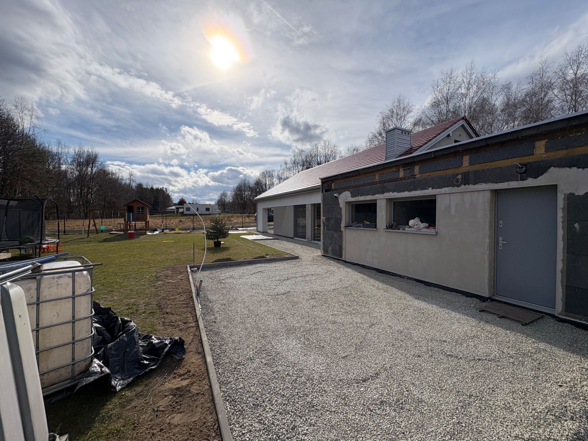 Utwardzony żwirem podjazd do domu w słoneczny dzień. Widoczne elementy konstrukcyjne budynku i fragment ogrodu z placem zabaw w tle.