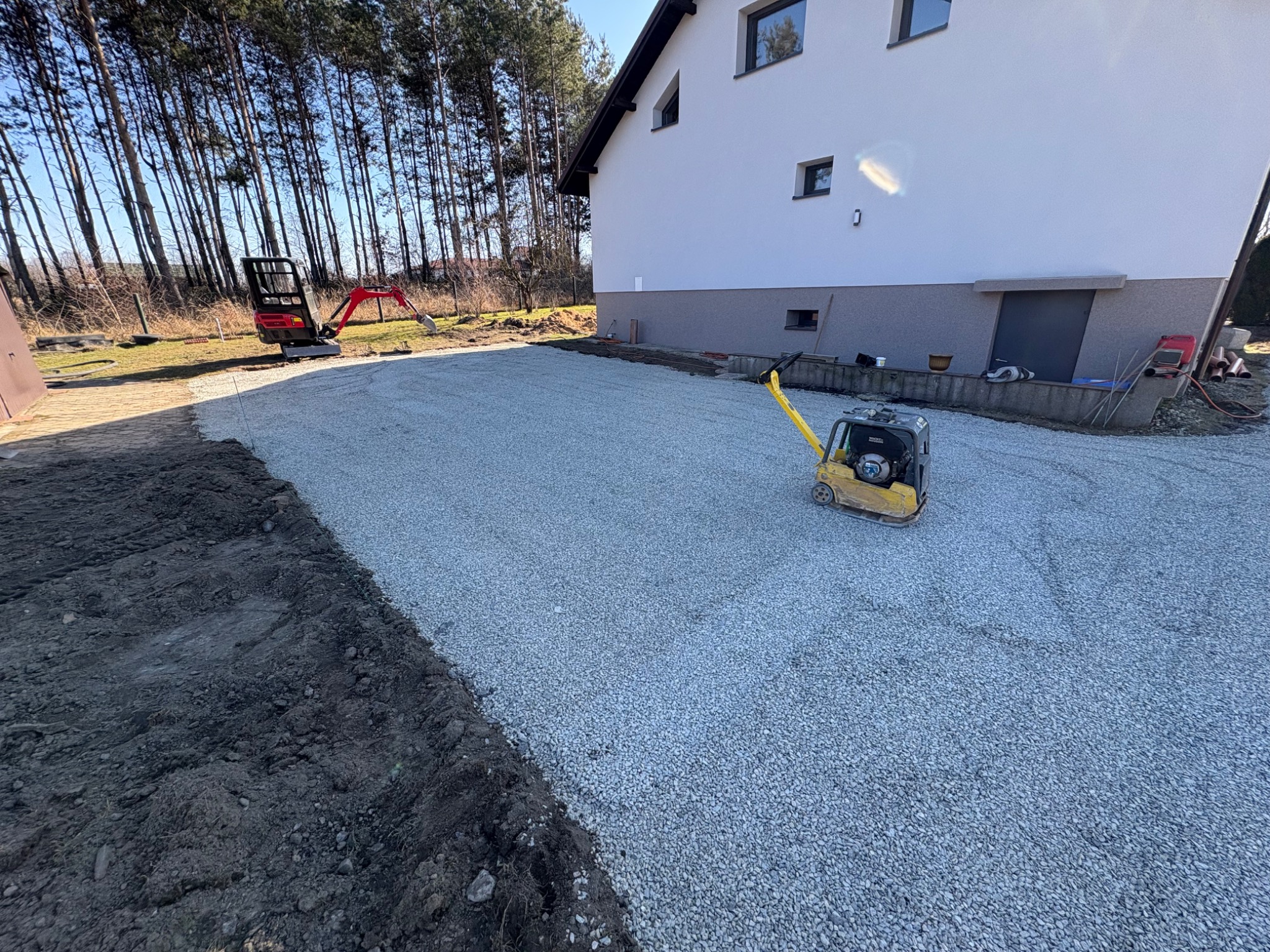 Przygotowany teren pod budowę: ubity żwir, widoczny zagęszczarką, w tle dom jednorodzinny i minikoparka. Grunt wyrównany, gotowy do dalszych prac.