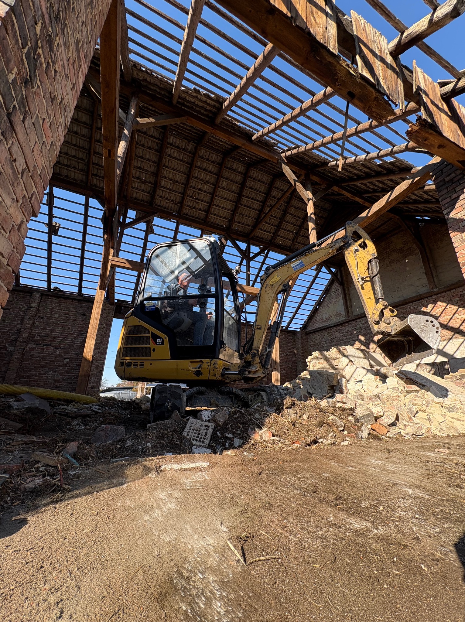 Żółta koparka CAT wewnątrz zrujnowanego budynku z cegły, częściowo zburzonego, z widocznym niebem przez uszkodzony dach. Gruz wewnątrz.