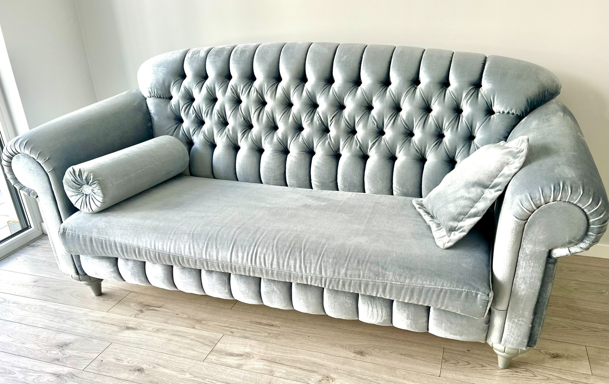 Elegancka, szara sofa Chesterfield z aksamitną tapicerką, ozdobnymi guzikami i poduszkami, ustawiona na drewnianej podłodze w jasnym wnętrzu. Widok z przodu.