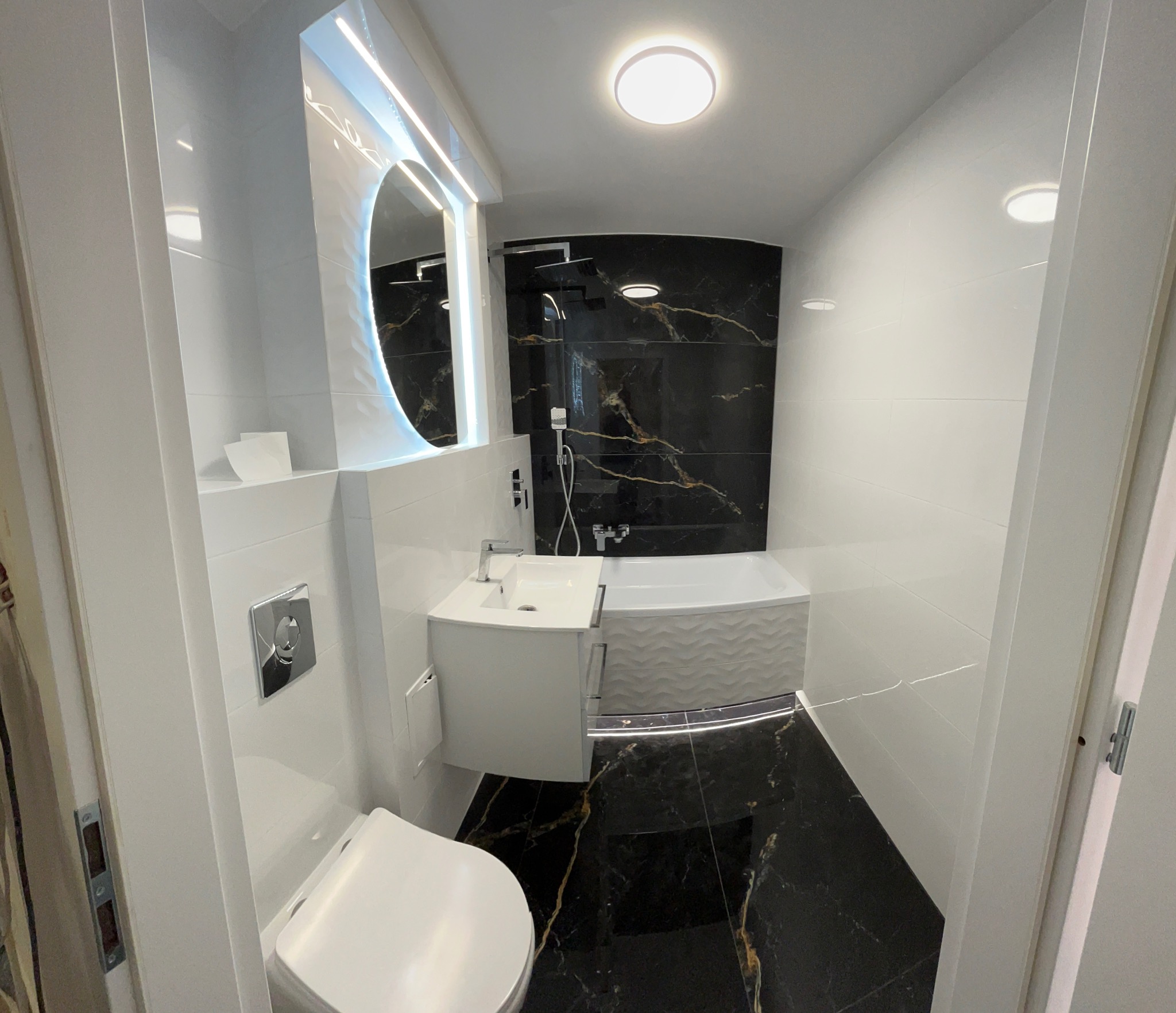 Nowoczesna łazienka z czarną marmurową ścianą za wanną, owalnym lustrem z podświetleniem LED, białą umywalką z szafką i podwieszaną toaletą, wykończona białymi płytkami i czarną marmurową podłogą...