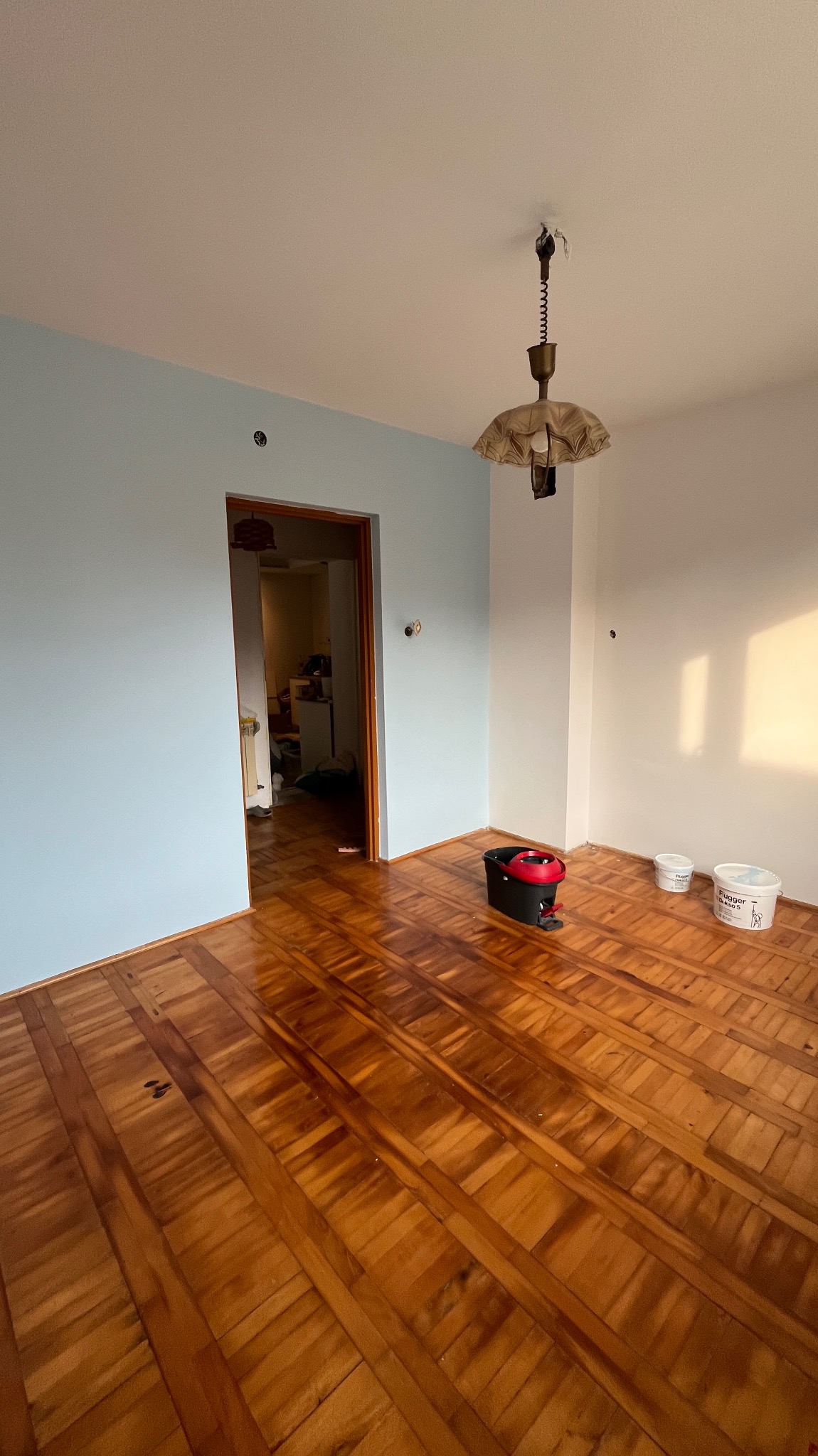 Pokój po częściowym malowaniu ścian na jasny błękit, widoczne dwa wiadra z farbą Flagger, mop z wyciskarką, parkiet, lampa wisząca i niedokończone gładzie na ścianie.