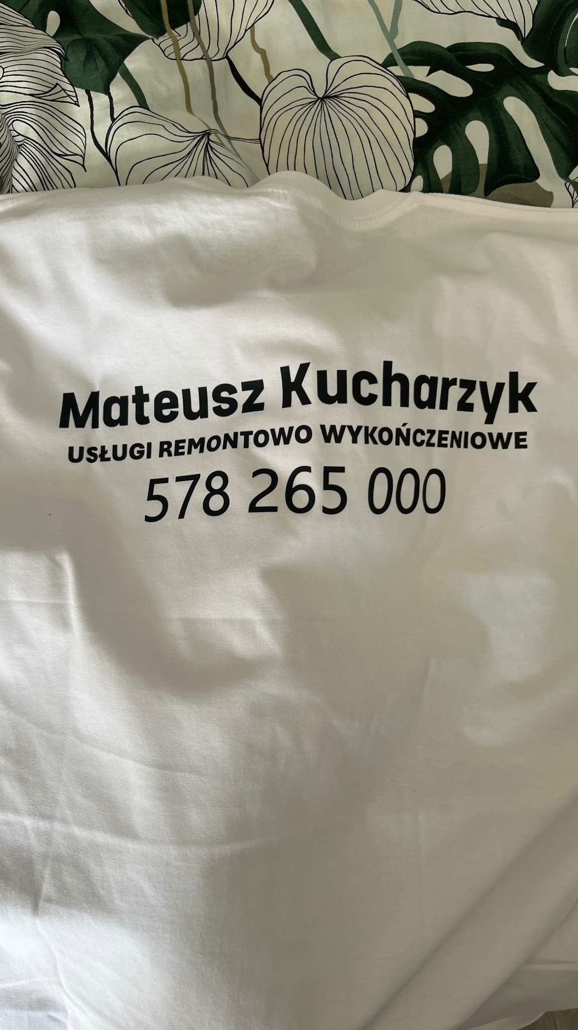 Biała koszulka z nadrukowanym imieniem i nazwiskiem Mateusz Kucharczyk, informacją o usługach remontowo-wykończeniowych oraz numerem telefonu 578 265 000, leżąca na pościeli w liście.