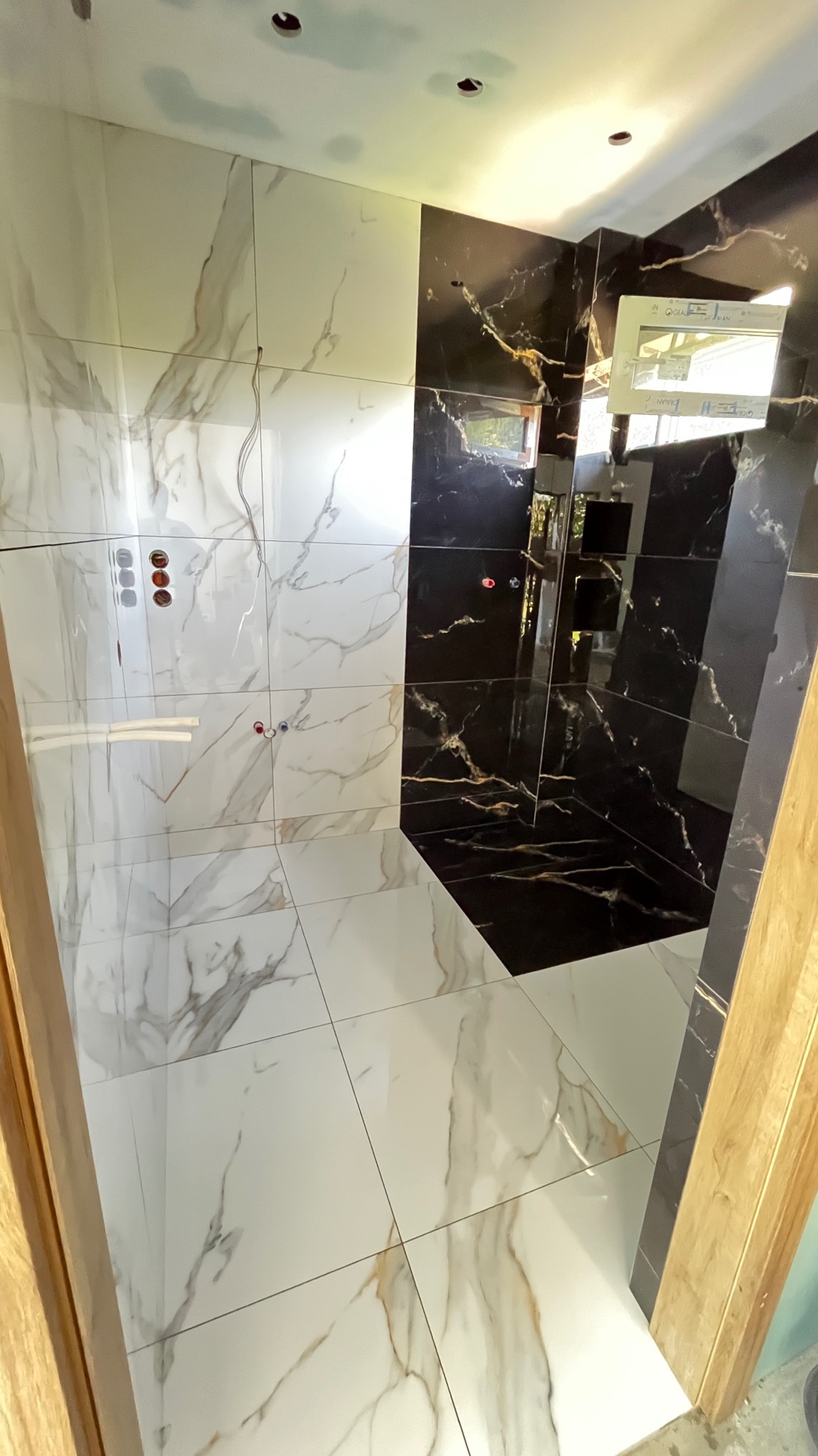 Wnętrze łazienki w trakcie wykańczania, z białymi i czarnymi płytkami imitującymi marmur, widoczne otwory na armaturę i wystające przewody, surowy sufit.