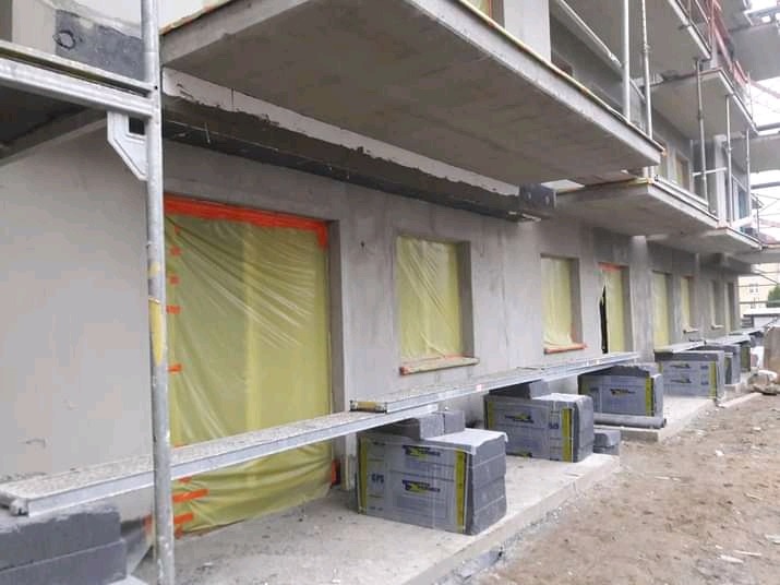 Budynek wielorodzinny w trakcie ocieplania elewacji, okna zabezpieczone folią, widoczne rusztowania i styropianowe płyty przygotowane do montażu.