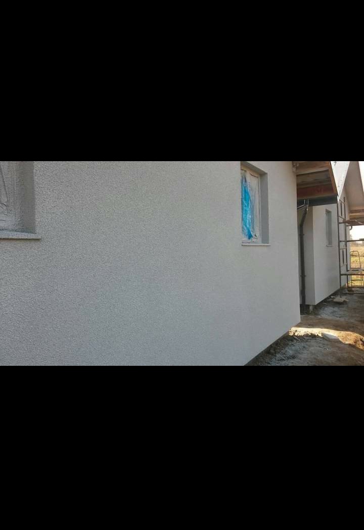 Świeżo otynkowana elewacja domu jednorodzinnego w jasnym odcieniu szarości; okno zabezpieczone folią malarską.