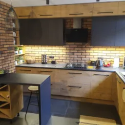 Nowoczesna kuchnia z ceglaną ścianą, drewnianymi i czarnymi szafkami, wyspą z barkiem na wino i czarnym blatem, oświetlona ciepłym światłem.