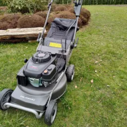 Szara kosiarka spalinowa marki Weibang stoi na trawniku z widocznymi liśćmi, w tle krzewy i drzewa iglaste.