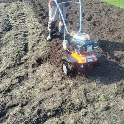 TOMASZ STARZAK GREENBI - Operator w roboczych spodniach i gumowcach obsługuje glebogryzarkę marki Stihl na polu z przygotowaną, spulchnioną glebą.
