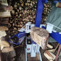 Hydrauliczna łuparka do drewna Cormak LUP 8E z rozłupywanym kawałkiem drewna na tle stosu pociętych klocków drewna.