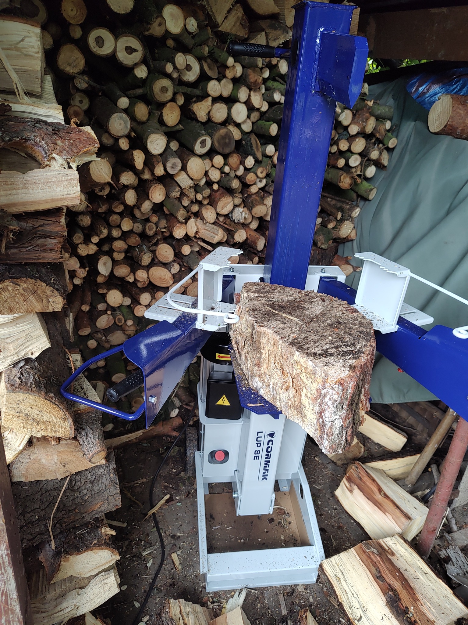 Hydrauliczna łuparka do drewna Cormak LUP 8E z rozłupywanym kawałkiem drewna na tle stosu pociętych klocków drewna.