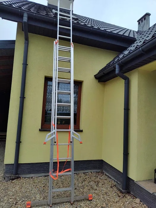 Rozstawiona, teleskopowa drabina aluminiowa oparta o dach domu jednorodzinnego, zabezpieczona pomarańczowymi pasami, widok z dołu.
