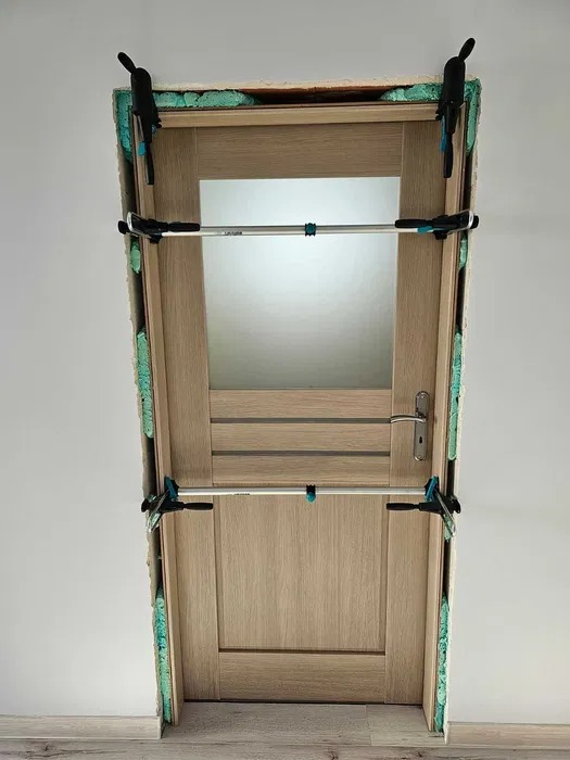 Montaż ościeżnicy drzwiowej z użyciem rozpieraka, widoczna piana montażowa i klamry mocujące.