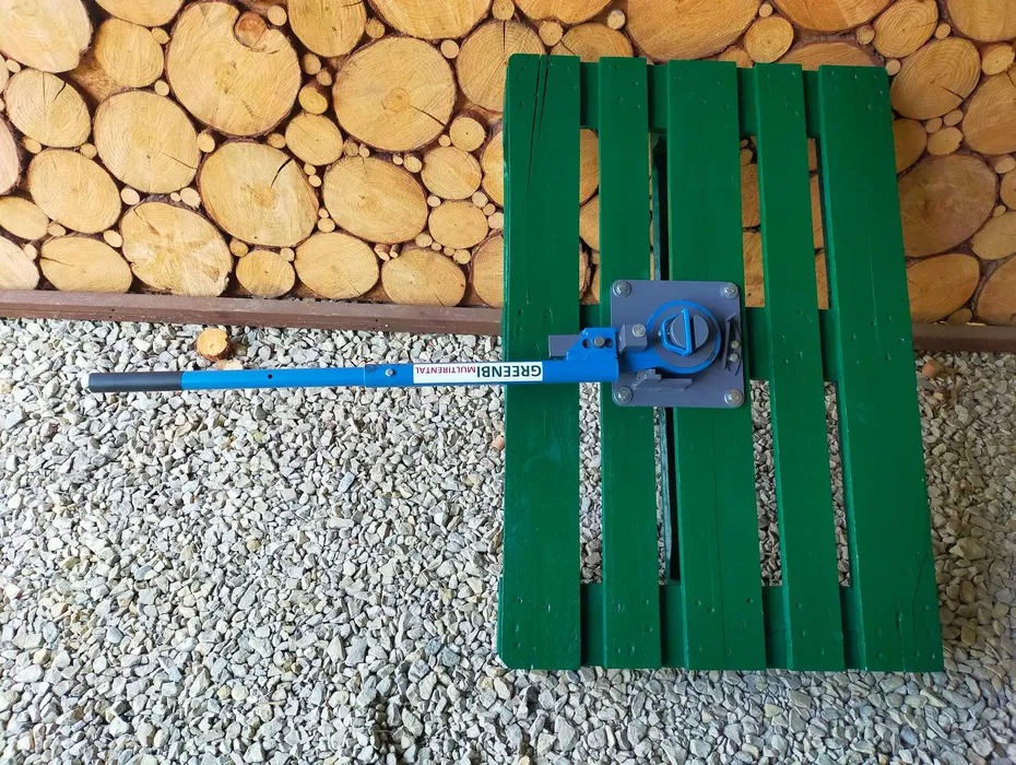 Giętarka do prętów marki Greenbi Multirental, leżąca na zielonej palecie na tle ściany z drewna i żwirowego podłoża.