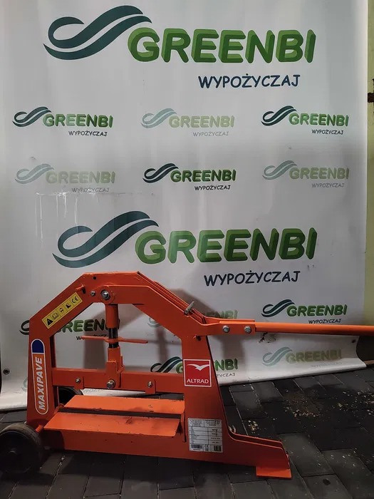 Pomarańczowa gilotyna do kostki brukowej marki Maxipave na tle ściany z logo firmy Greenbi wypożyczającej sprzęt.
