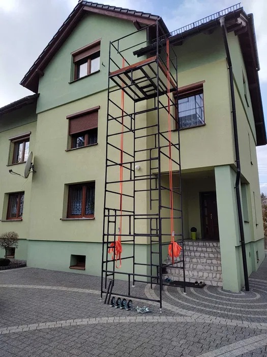 Rozstawione rusztowanie warszawskie przed dwukondygnacyjnym domem z zieloną elewacją, widoczne elementy montażowe na kostce brukowej.