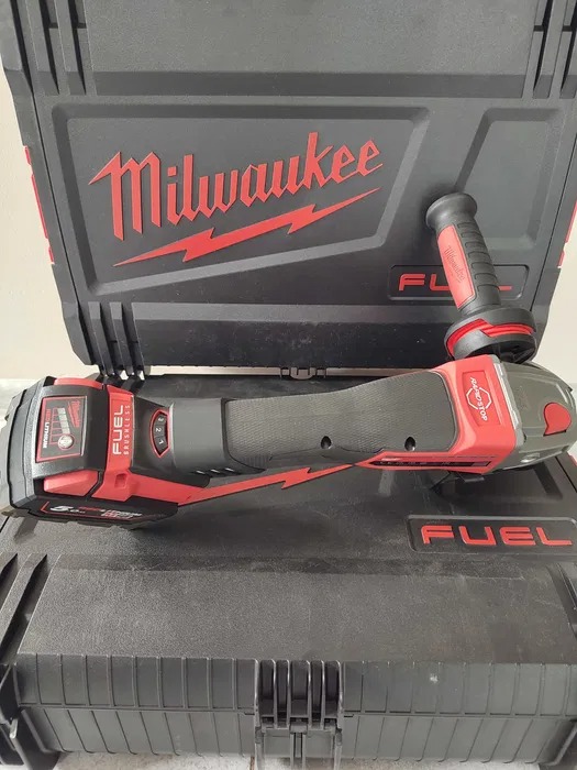 Akumulatorowa szlifierka kątowa Milwaukee FUEL z dodatkowym uchwytem, leżąca na czarnej skrzynce z czerwonym logo Milwaukee.