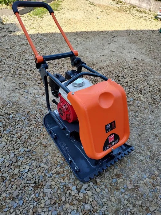 Pomarańczowo-czarna zagęszczarka gruntu Belle PCX 20/50 z silnikiem Honda, stojąca na żwirowym podłożu.