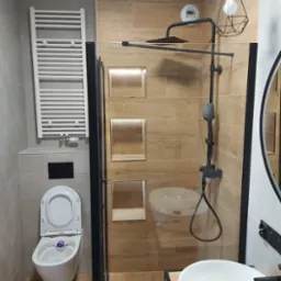 Nowoczesna łazienka z drewnopodobnymi płytkami, kabiną prysznicową typu walk-in z czarnymi profilami, wiszącą toaletą i grzejnikiem drabinkowym, oświetlona geometryczną lampą wiszącą.