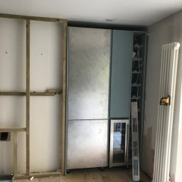 Made - Wnęka w trakcie zabudowy: szkielet z drewnianych belek przy ścianie, obok moduł szafy wnękowej z chłodziarką na wino i półkami na butelki, widoczny poziomica laserowa na grzejniku.