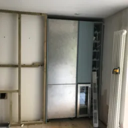 Wnęka w trakcie zabudowy: szkielet z drewnianych belek przy ścianie, obok moduł szafy wnękowej z chłodziarką na wino i półkami na butelki, widoczny poziomica laserowa na grzejniku.