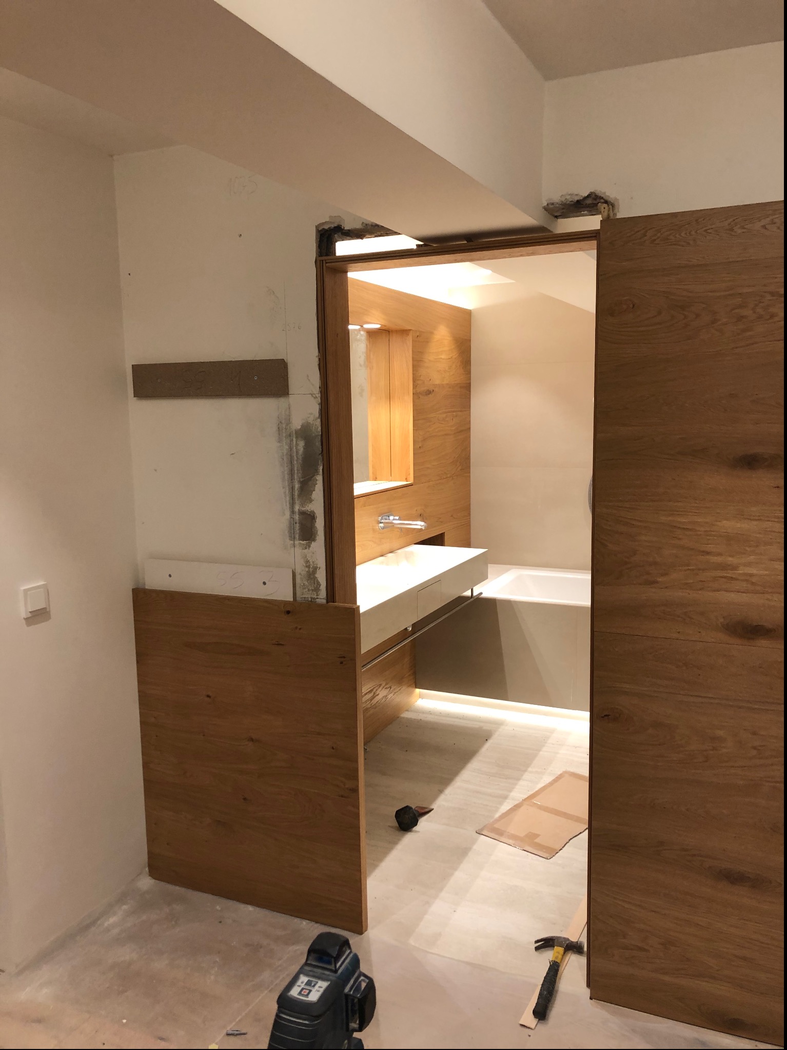 Łazienka w trakcie remontu z drewnianą zabudową, widoczna wanna, umywalka, laser krzyżowy na podłodze i młotek, surowe wykończenie ścian.