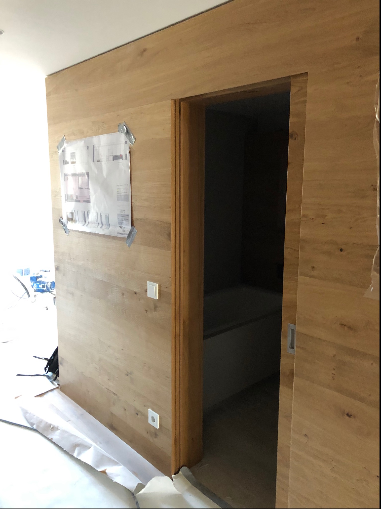 Ściana wykończona drewnianymi panelami z ukrytymi drzwiami przesuwnymi do łazienki, na ścianie przyklejony plan pomieszczenia, podłoga zabezpieczona folią ochronną.